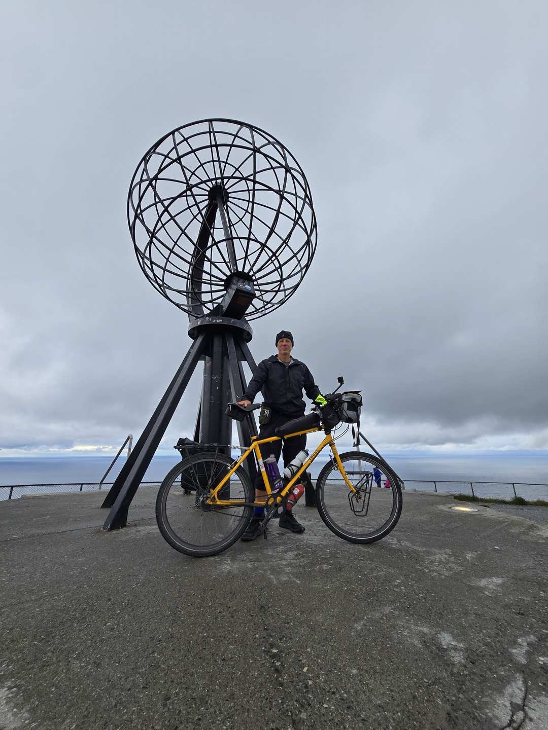 Cyclist standing with a loaded touring bicycle under the iconic Nordkapp globe sculpture on the cliff top